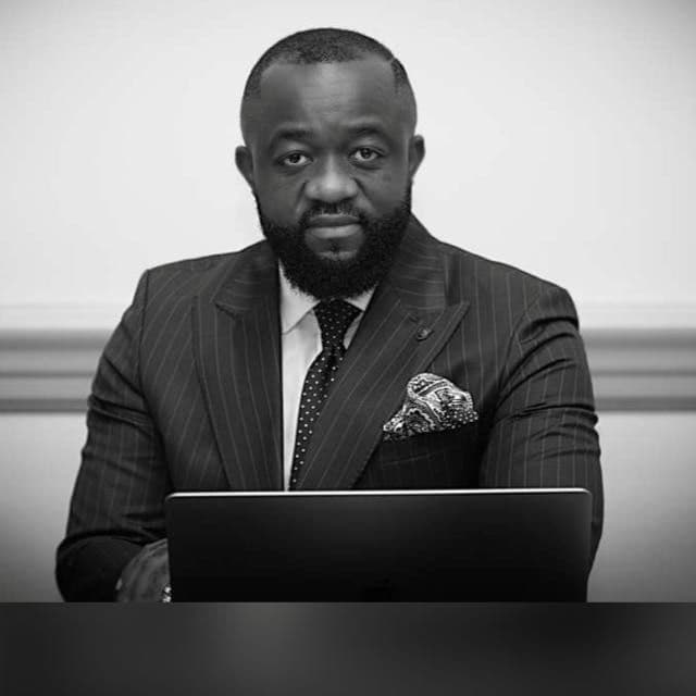 Bearded man in a pinstripe suit and tie sits behind a laptop in monochrome portrait.